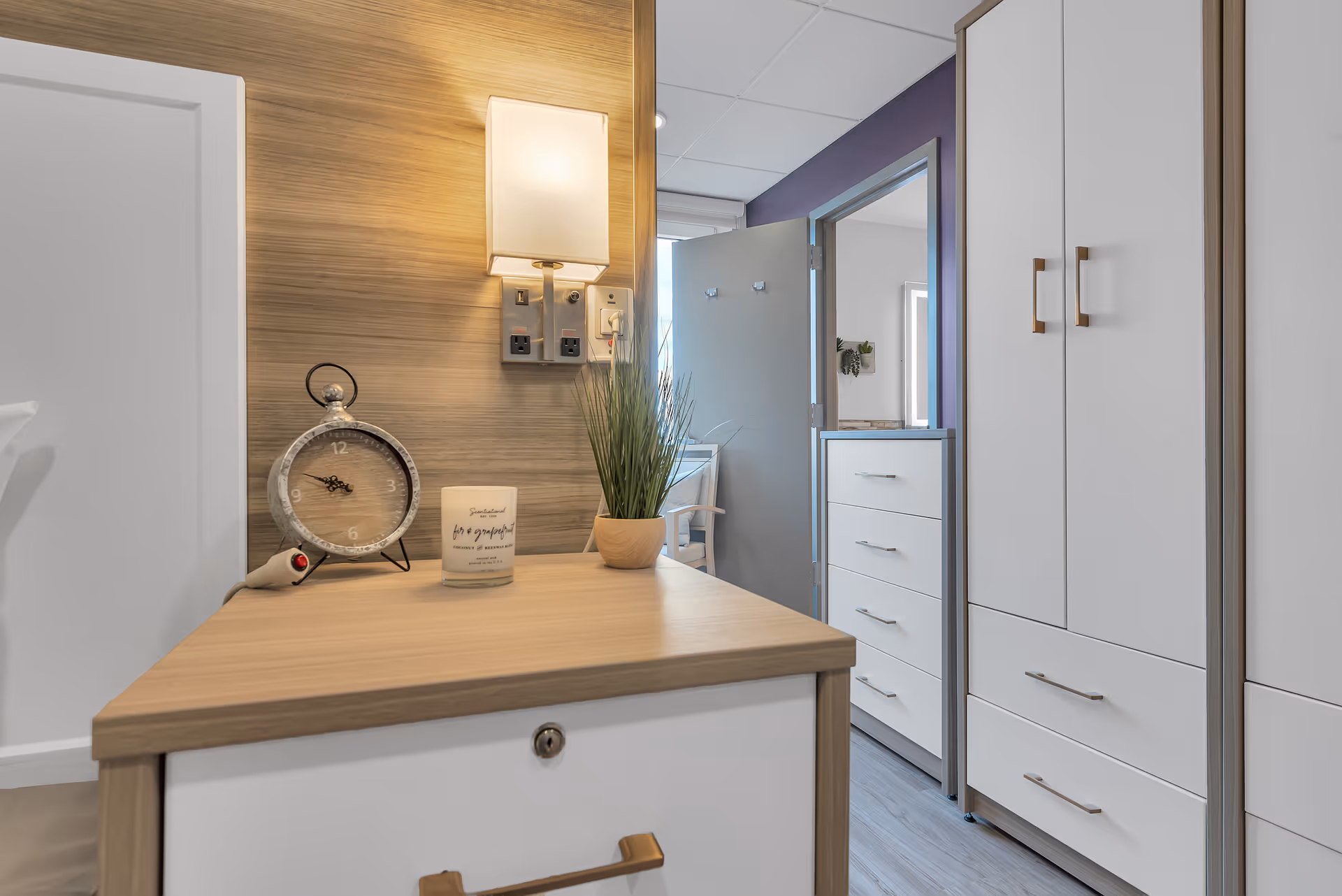A corner of a bedroom in The Veranda Assisted Living facility showing a wooden nightstand with a vintage-style clock, a candle, and a small potted plant. A wall-mounted light fixture is above the nightstand. In the background, there is a partially open door leading to another room with white cabinets and a purple accent wall.