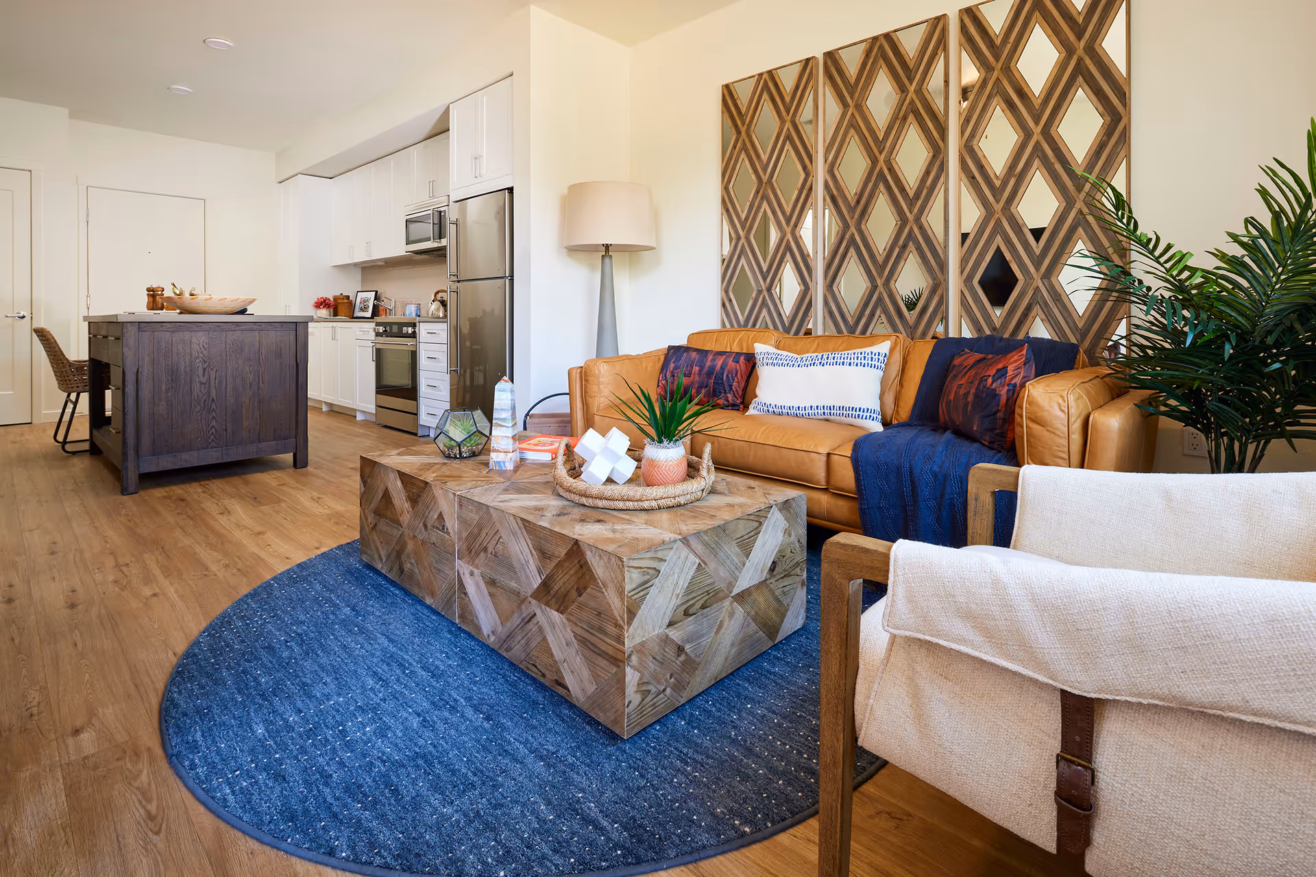 Open-plan living area with a tan leather sofa, wooden geometric coffee table on a blue rug, and a kitchen island in the background.