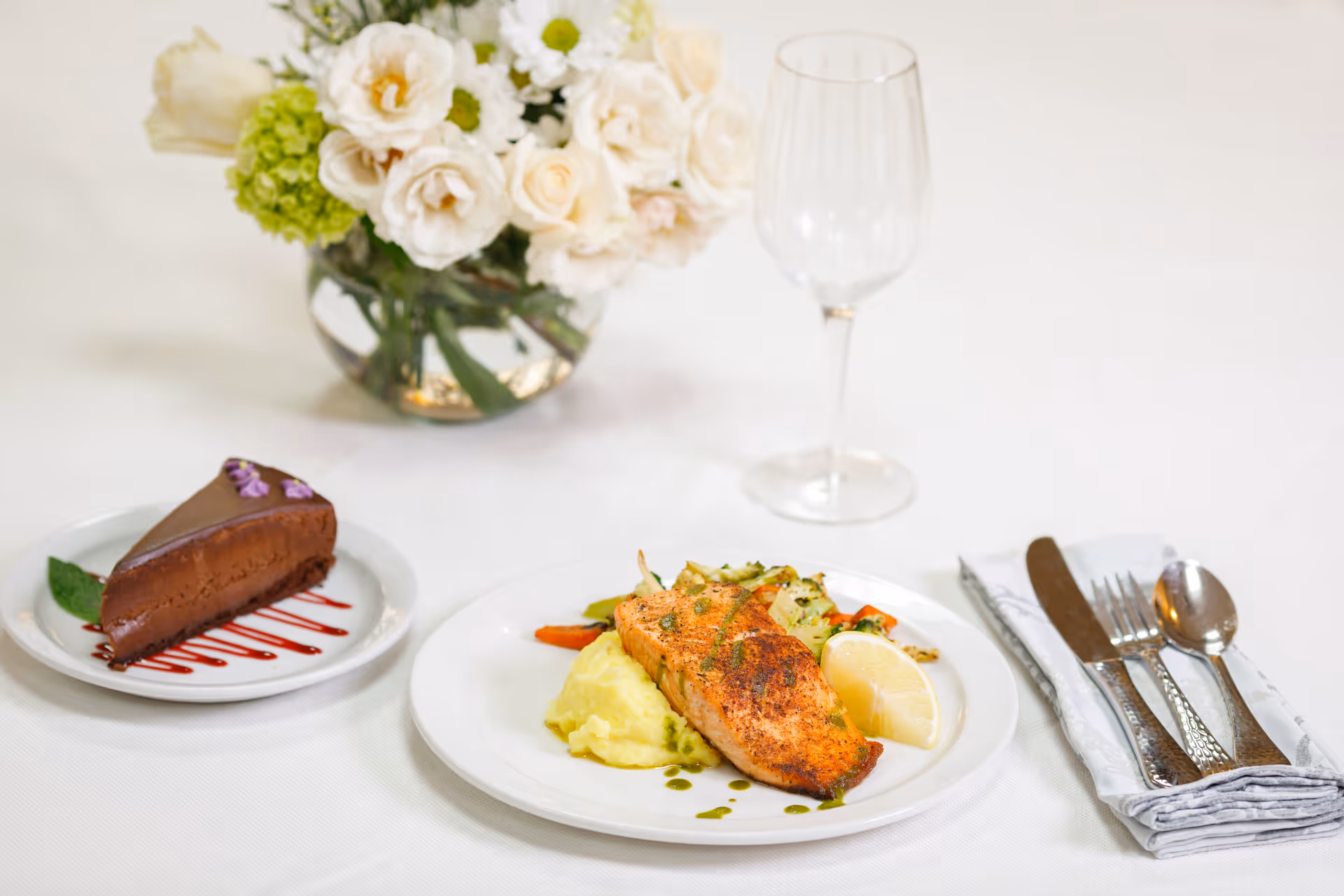 A plated meal featuring a piece of seasoned salmon on mashed potatoes with mixed vegetables and a lemon wedge, accompanied by a slice of chocolate cake with raspberry sauce on a separate plate. There is a wine glass and a neatly folded napkin with a knife, fork, and spoon on a white tablecloth. A vase with white and green flowers is in the background.