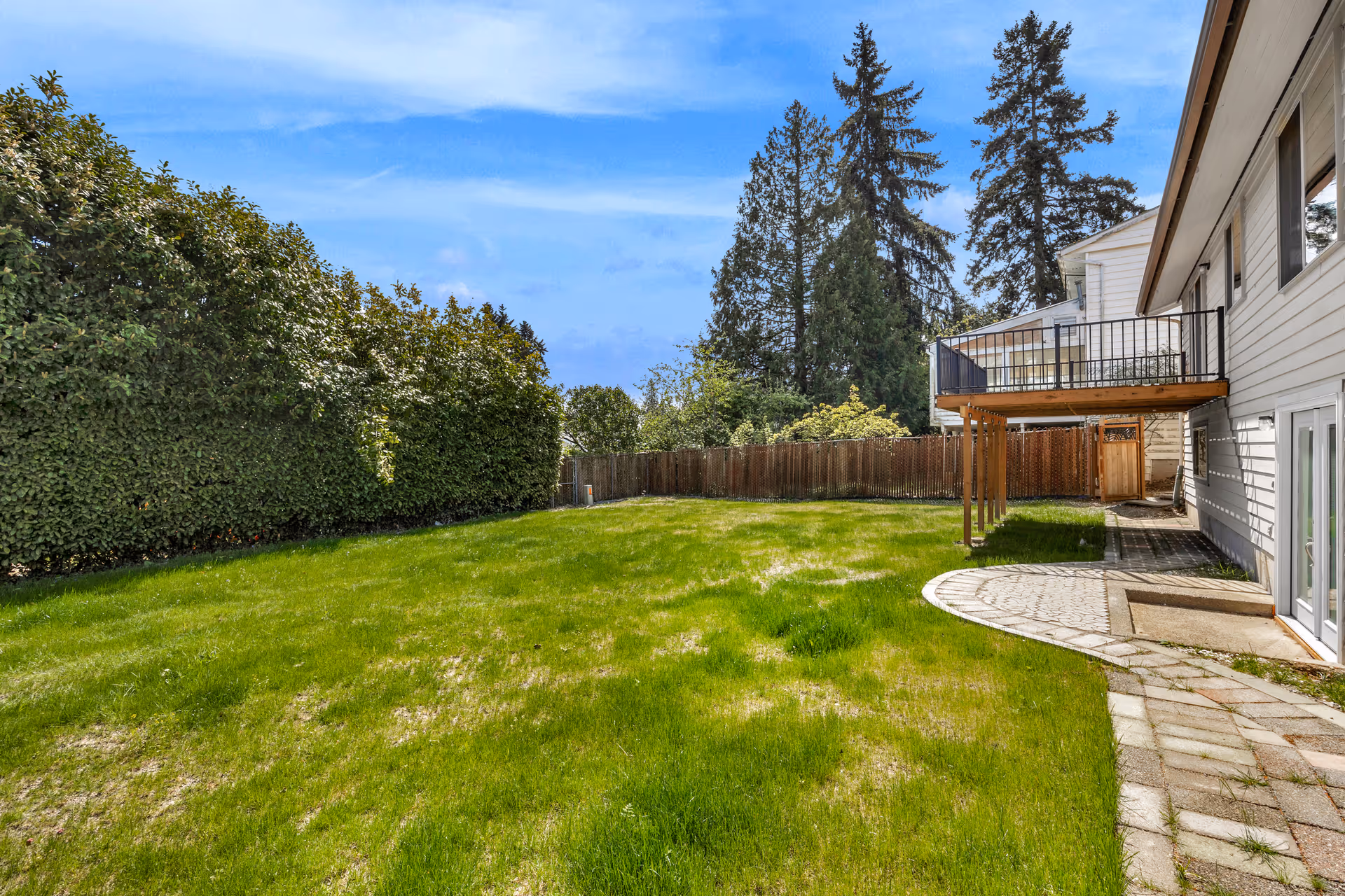 A spacious backyard with green grass, a paved walkway, and a wooden deck attached to a white house. Tall trees and a wooden fence surround the yard under a blue sky with some clouds.