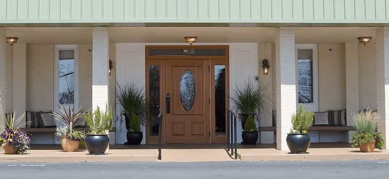 Front entrance of a building with a wooden door featuring an oval glass panel, sidelights, potted plants, benches, and a covered porch.