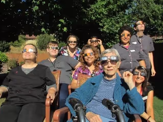 A group of eight people, mostly elderly women, sitting and standing outdoors on a sunny day wearing special glasses, possibly for viewing a solar eclipse. They are smiling and appear to be enjoying the event in a garden or patio area with trees and greenery in the background.