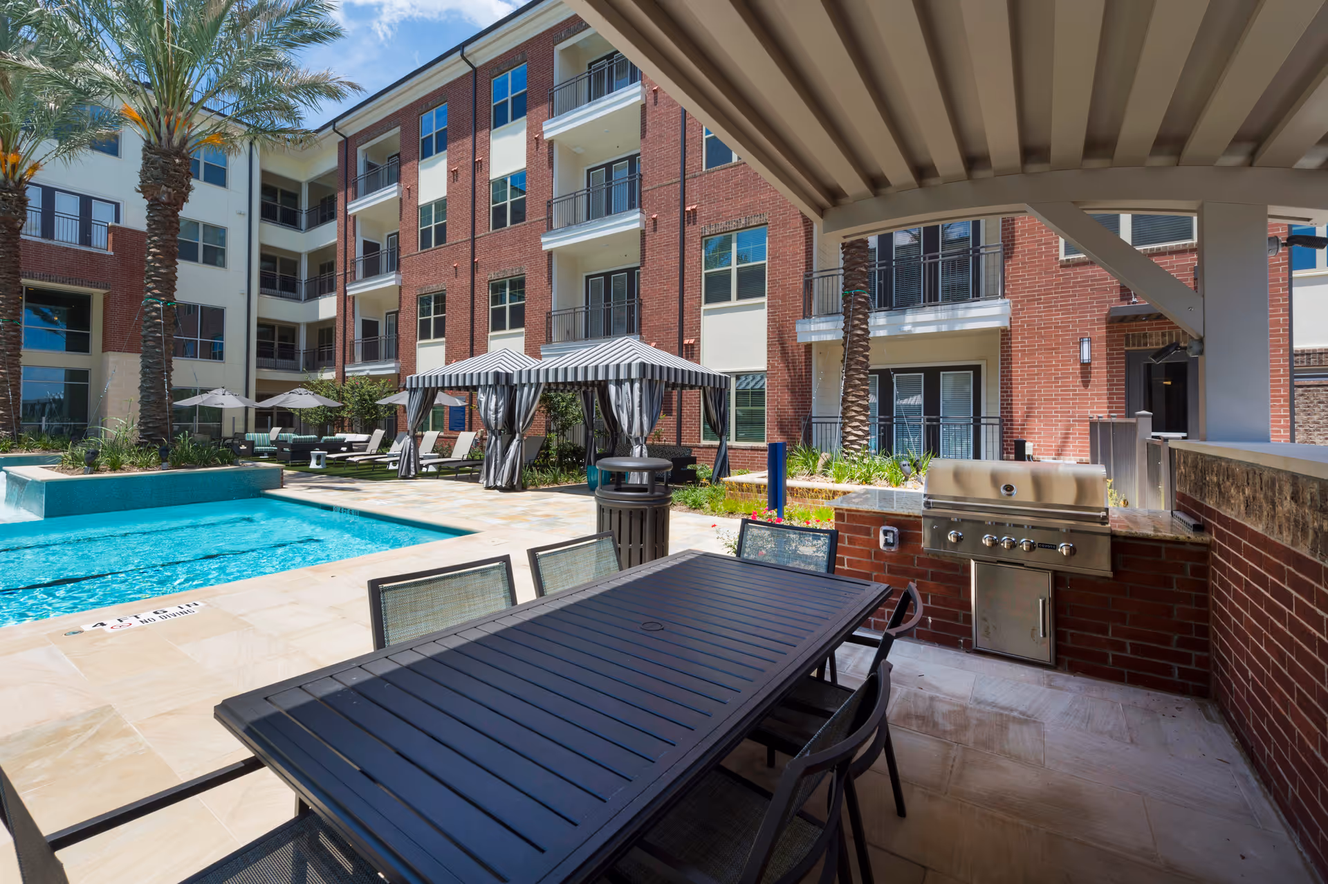 Outdoor courtyard with a swimming pool, striped cabanas, lounge chairs, dining table and built-in grill in front of a multi-story brick residential building.