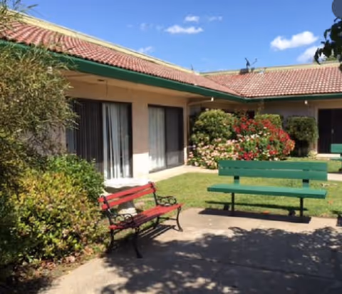 Outdoor courtyard area of a senior living facility with two benches, one red and one green, surrounded by bushes and flowering plants. The building has beige walls and a red tiled roof under a blue sky with a few clouds.