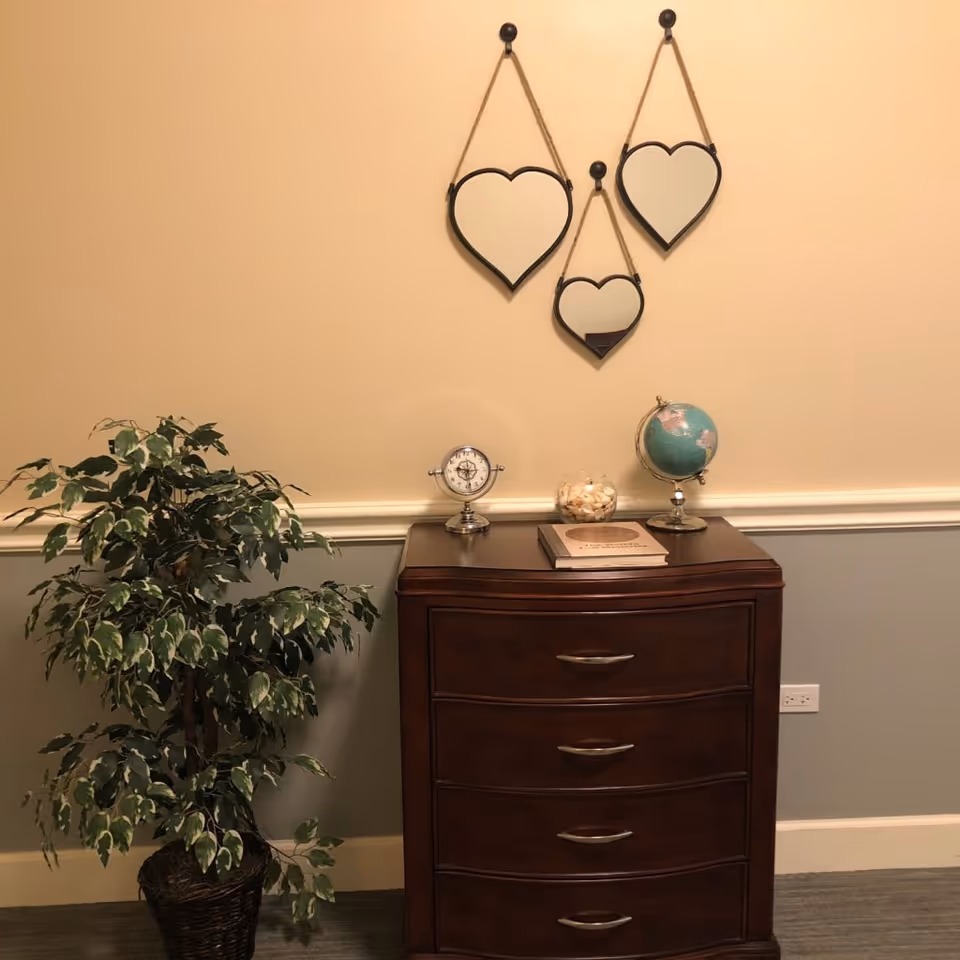 A wooden chest of drawers with a small globe, a clock, a glass bowl filled with shells, and a book on top. Above the chest, three heart-shaped mirrors hang on the wall. To the left of the chest is a potted leafy plant. The wall is painted beige on the upper half and gray on the lower half with a white chair rail separating the two colors.