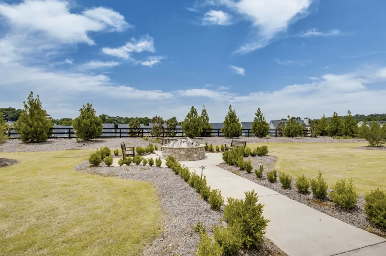 Outdoor garden area with a circular stone fire pit surrounded by wooden benches and small bushes. A concrete pathway leads to the fire pit, with neatly trimmed grass and trees in the background under a partly cloudy blue sky.