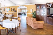 A cozy interior common area with round tables covered with white tablecloths and wooden chairs. There is a patterned sofa facing a wooden entertainment center with a television and glass cabinets. The room has wooden flooring and beige walls with framed pictures. An arched doorway leads to another room with more tables and chairs.