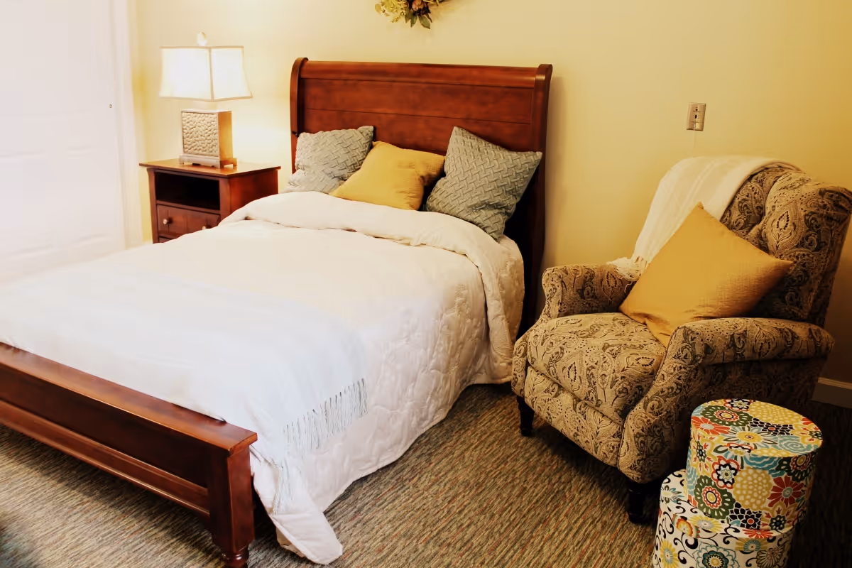 A cozy bedroom with a wooden bed frame, white bedding, and three pillows in yellow and gray. Next to the bed is a wooden nightstand with a lamp. There is a patterned armchair with a yellow pillow and a white throw blanket, and two colorful floral-patterned round storage boxes on the floor beside the chair.