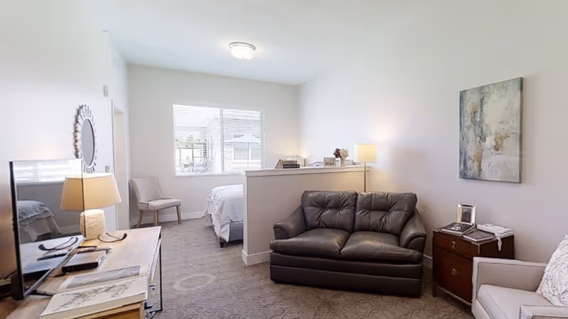 A cozy assisted living room with a black leather loveseat, a beige armchair, a wooden side table with a lamp and picture frame, a TV on a wooden stand, and a bed partially visible behind a half wall under a window with blinds.