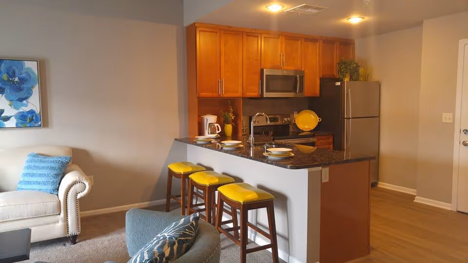 Interior view of a senior living apartment showing a kitchen with wooden cabinets, a stainless steel microwave and refrigerator, a black countertop island with three yellow cushioned bar stools, and a living area with a beige armchair and a blue pillow. A blue floral painting is hung on the wall.
