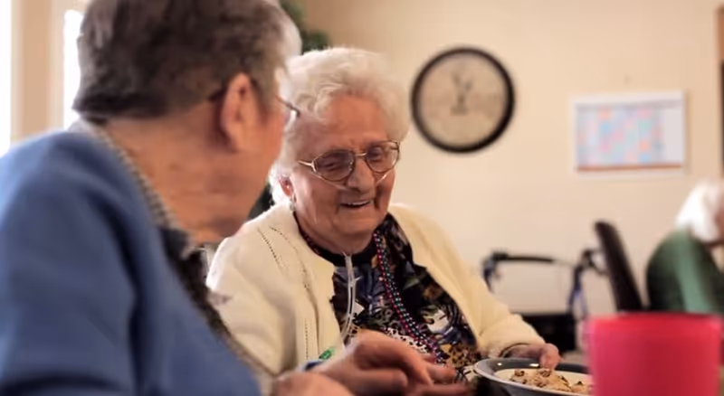 Two elderly women sit at a table smiling while sharing a meal in a dining area.