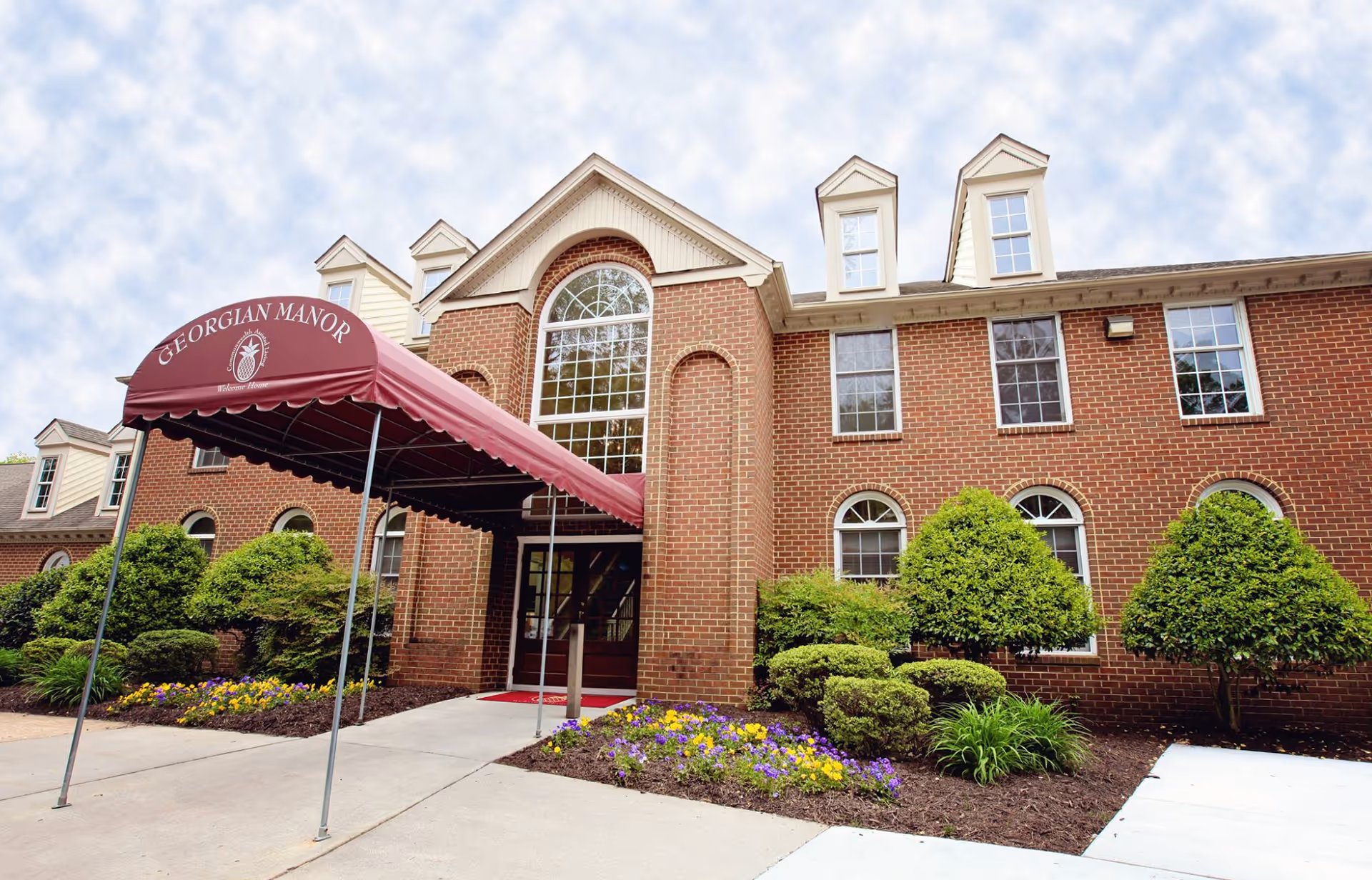 Brick-front senior living building entrance with a maroon 'GEORGIAN MANOR' awning, arched windows, and landscaped flowerbeds.