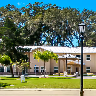 Two-story beige senior living building with a covered entrance, palm trees, a lawn and a lamp post under a bright blue sky.