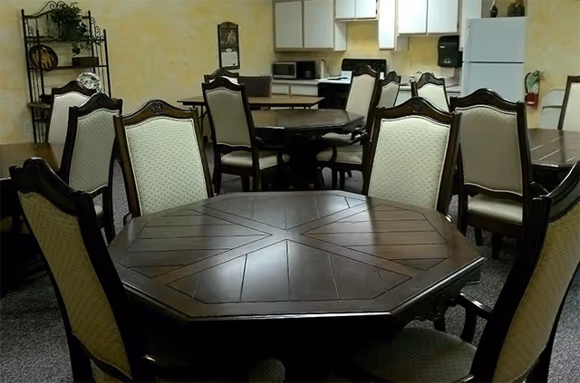 Interior room with multiple dark wooden tables and cushioned chairs arranged around them. The background shows white cabinets, a microwave, and a paper towel dispenser mounted on the wall. The walls are painted yellow with some decorative items and a plant on a metal shelf.