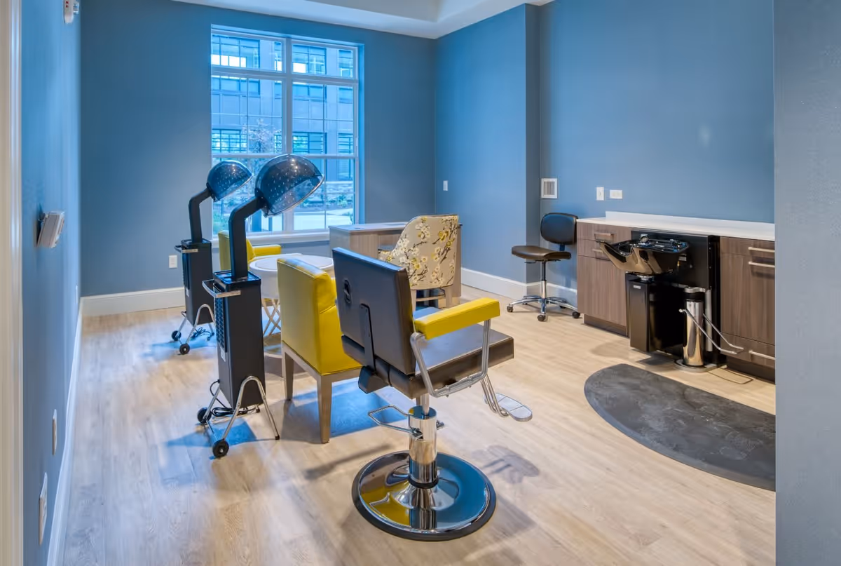 Interior view of a salon room with two yellow salon chairs, two hair dryer stations, a floral armchair, a black swivel chair, and a hair washing station against a blue wall with a large window letting in natural light.