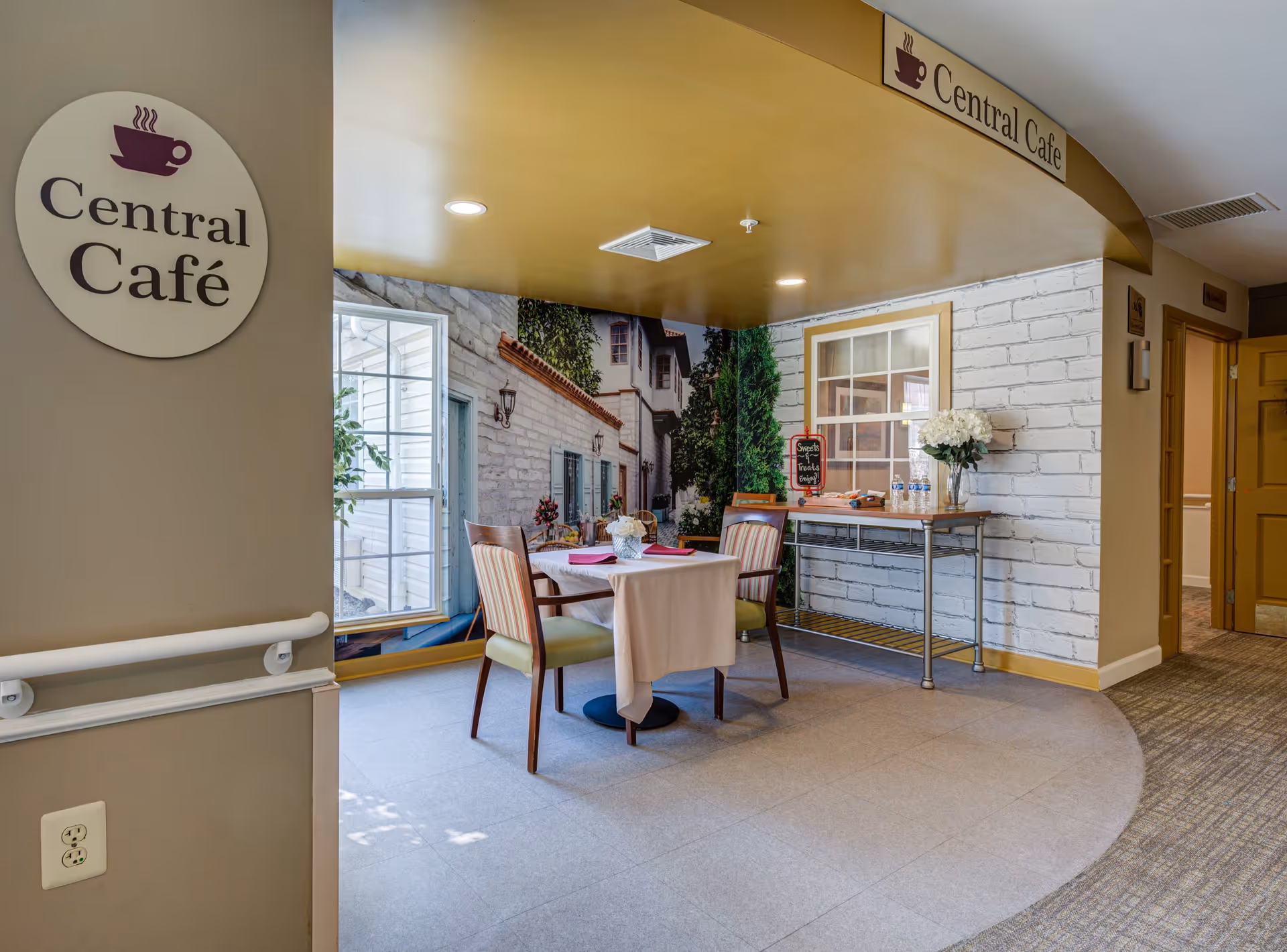 A small café area inside a senior living facility with a sign reading 'Central Café'. The space includes a table covered with a white tablecloth, two chairs with striped upholstery, a side table with water bottles and a vase of white flowers, and a wall mural depicting a quaint outdoor scene with windows and greenery.
