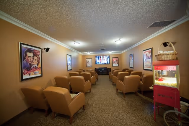 A small movie theater room with two rows of tan leather armchairs facing a flat-screen TV mounted on the far wall. The walls are decorated with framed classic movie posters, including Casablanca. There is a red popcorn machine with a basket on top on the right side of the room.
