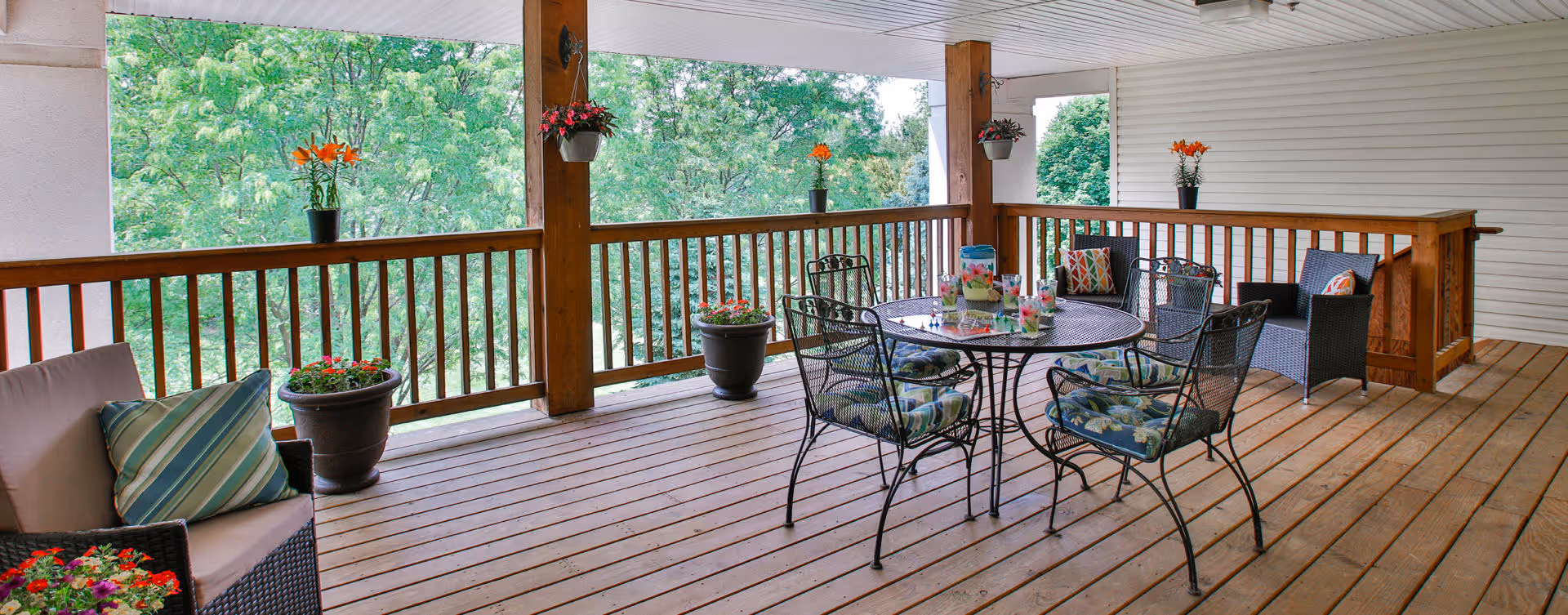 Covered wooden deck with outdoor seating including a round metal table with four cushioned chairs, two cushioned armchairs, and several potted plants with flowers. The deck overlooks green trees in the background.