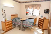 A single bedroom in a senior living facility with a hospital-style bed covered in a floral bedspread, a bedside table with a lamp and decorative items, a wooden dresser, a wall-mounted television, and a window with curtains letting in natural light.