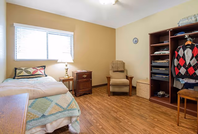 A cozy bedroom with a single bed covered with a quilt and pillows, a wooden nightstand with a lamp, a comfortable armchair with a blanket draped over the back, a wooden open wardrobe with clothes hanging and folded, and a small wooden table. The room has wood flooring and beige walls with a window letting in natural light.
