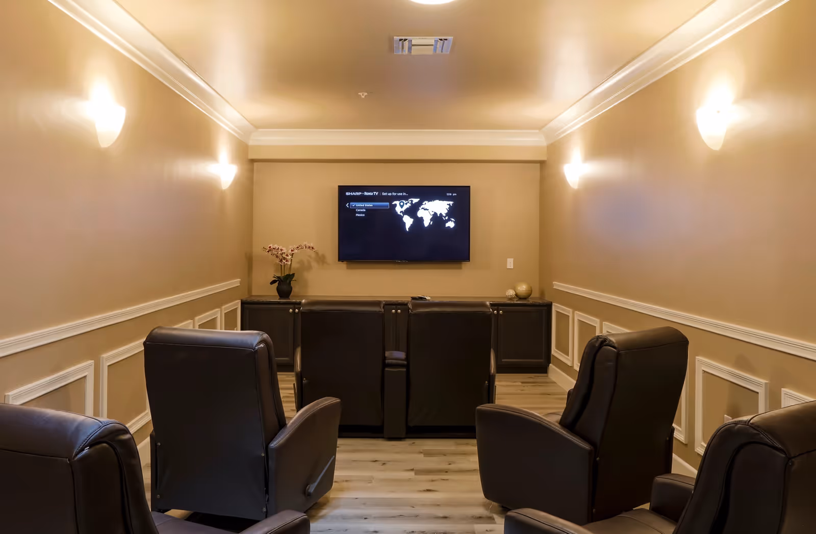A small home theater room with six black leather recliner chairs arranged in two rows facing a wall-mounted flat-screen TV. The walls are beige with white trim and decorative molding, and there are wall sconces providing warm lighting. A cabinet below the TV holds a potted orchid and decorative spheres.