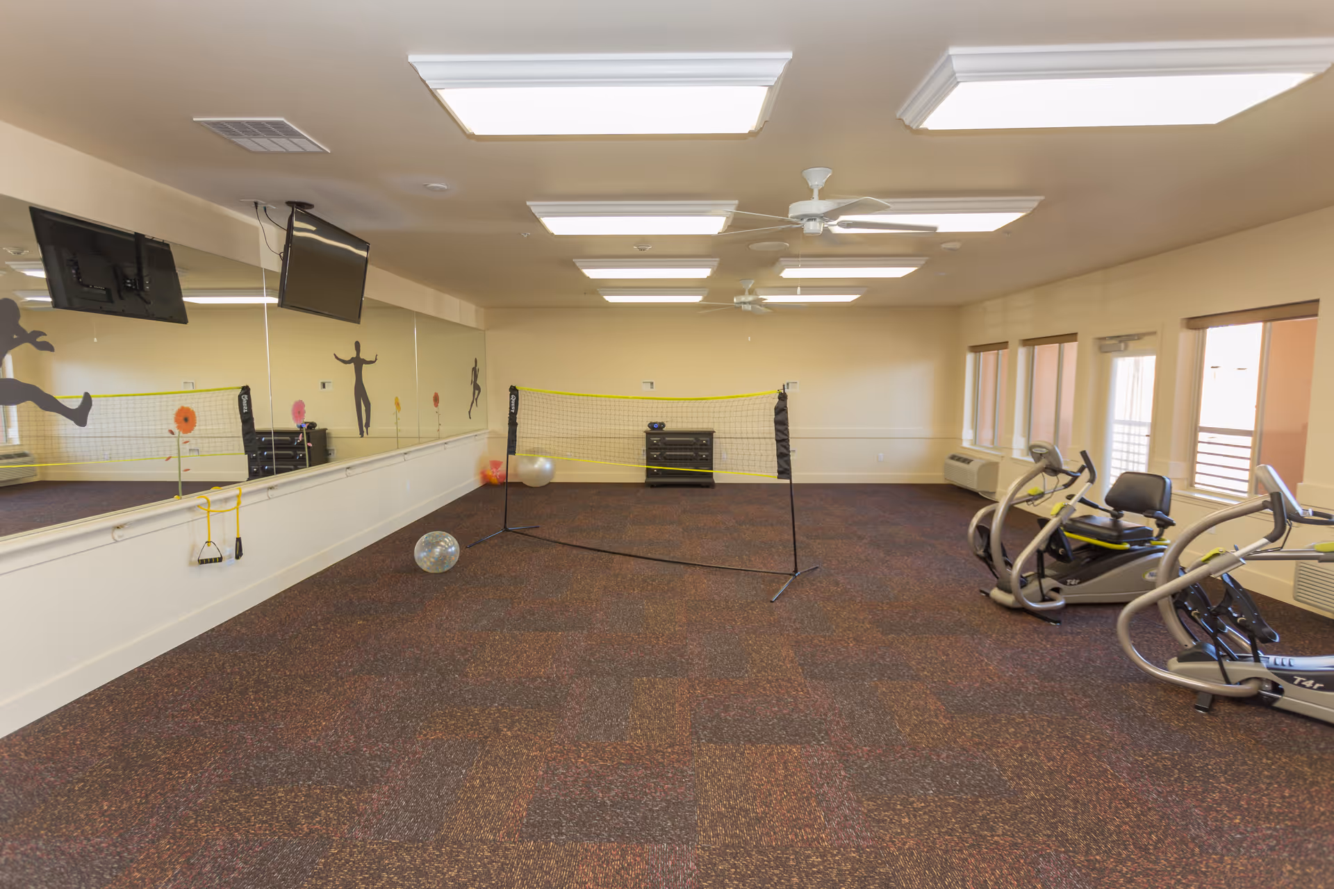 A spacious indoor exercise room with a carpeted floor, a small badminton net set up in the center, exercise balls on the floor, two stationary exercise bikes on the right side, large mirrors on the left wall with silhouettes of people in active poses, and multiple ceiling lights and fans. Windows on the right wall allow natural light into the room.