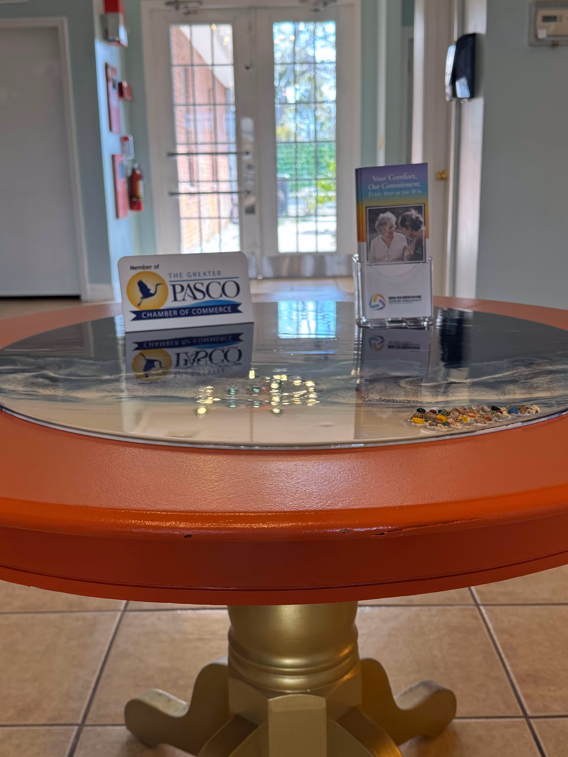 A round table with an orange edge and a golden pedestal base is positioned indoors on a tiled floor. On the table are two display stands: one with a sign indicating membership in The Greater Pasco Chamber of Commerce, and the other holding brochures from Braybrook Residence, Inc. In the background, there is a glass door letting in natural light and a wall-mounted phone on the right side.