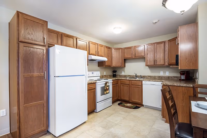 A clean kitchen with wooden cabinets, a white refrigerator, white stove with oven, white dishwasher, and a small dining table with two chairs. The countertops are brown granite, and the floor is tiled in a light beige color. The kitchen is well-lit with ceiling lights.