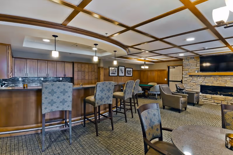 A senior living facility common area featuring a bar with high chairs, a stone fireplace with a mounted TV above it, comfortable armchairs, a round table with chairs, and framed pictures on the wall. The ceiling has a decorative grid pattern with recessed lighting.