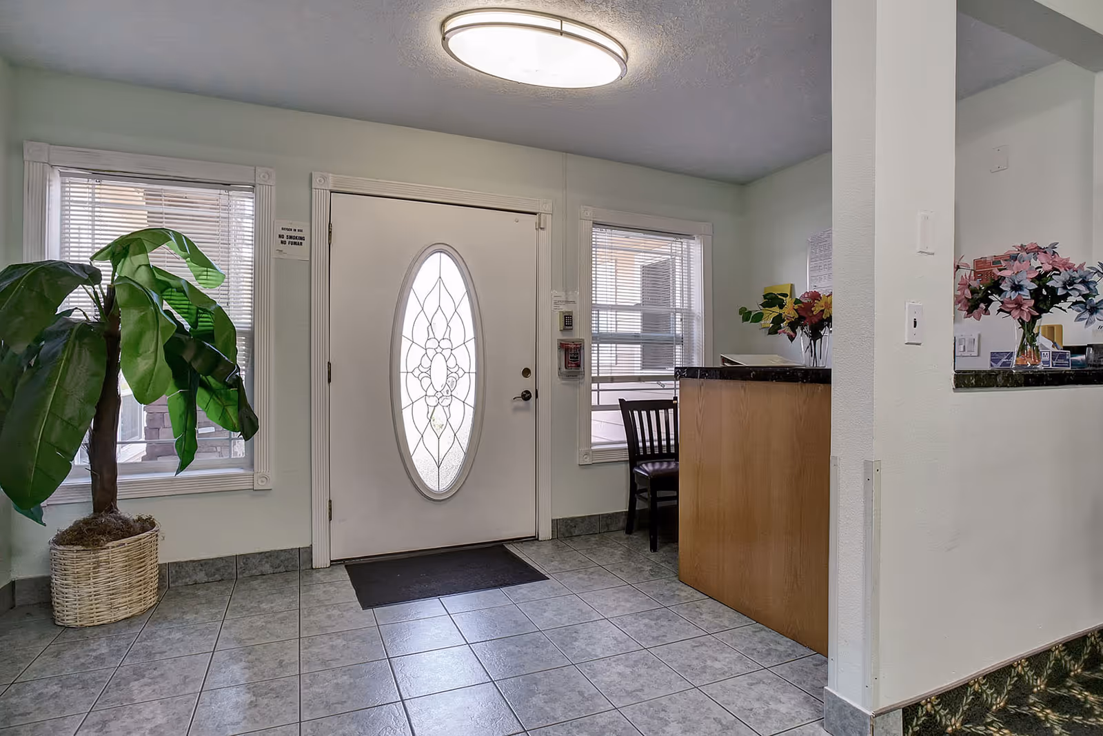 Entrance area of a facility with a white door featuring an oval decorative glass window, two windows with blinds on either side, a large potted plant to the left, and a wooden reception desk with a chair and flower arrangements to the right.