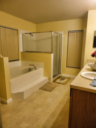 A bathroom with a built-in bathtub, a separate glass-enclosed shower, two windows with vertical blinds, a double sink vanity with a countertop, and a small rug on the floor.