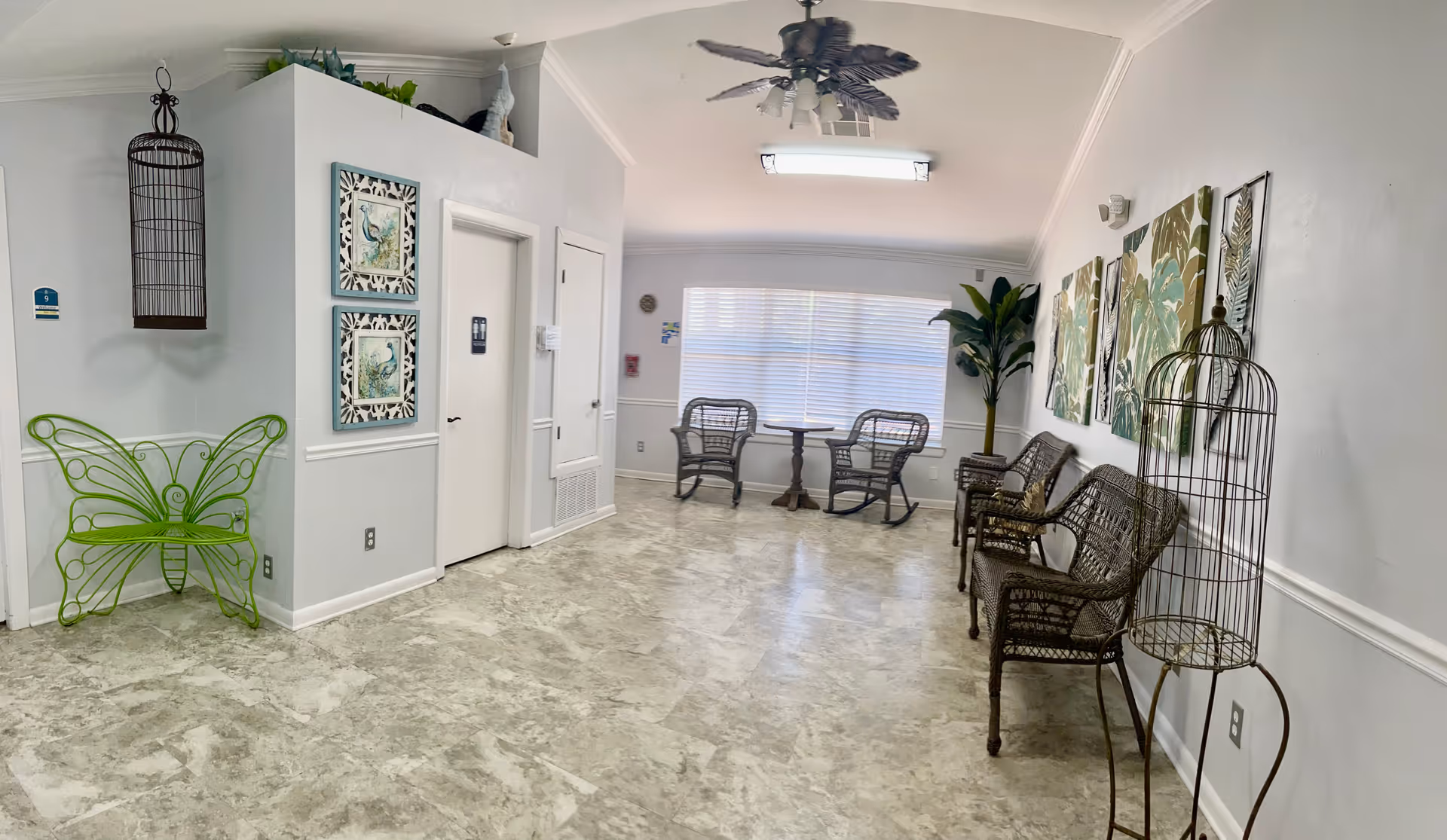 A bright and spacious waiting or sitting area with a light-colored tiled floor and white walls. The room features several wicker chairs arranged along the right wall, decorative birdcages, framed botanical artwork, a green butterfly-shaped metal bench, and a ceiling fan with leaf-shaped blades. A large window with blinds allows natural light to fill the space, and a small round table with two wicker chairs is positioned near the window. There are two closed doors on the left wall.