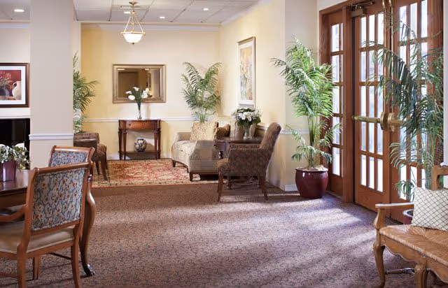 A well-lit senior living facility common area with patterned carpet, several upholstered chairs and sofas, potted plants, framed artwork on the walls, a wooden table with a vase of flowers, and large wooden doors with glass panels allowing natural light to enter.