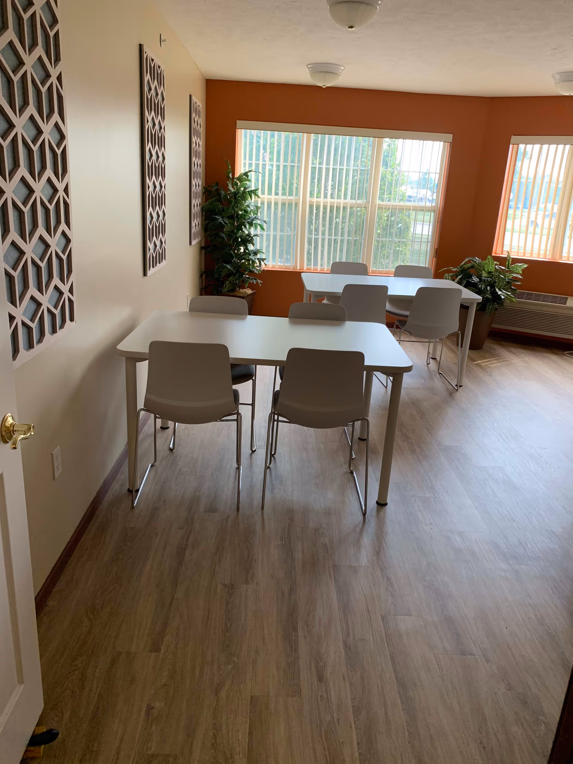A bright room with two white tables and six white chairs arranged around them. The room has light wood flooring, orange accent walls with large windows covered by vertical blinds, and decorative wall panels. There are two potted plants near the windows and a heating/cooling unit beneath one window.