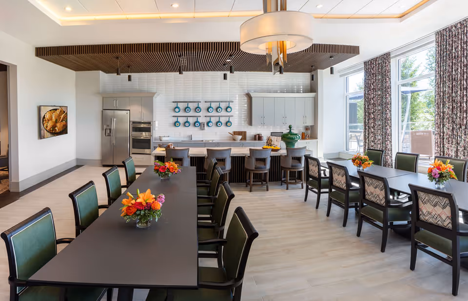 A bright dining room with two long black tables surrounded by green cushioned chairs. Each table has a floral centerpiece with orange and pink flowers. The room features a modern kitchen area in the background with white cabinets, stainless steel appliances, and a row of blue pans hanging on the wall. Large windows with floral curtains let in natural light, and a large circular light fixture hangs from the ceiling.
