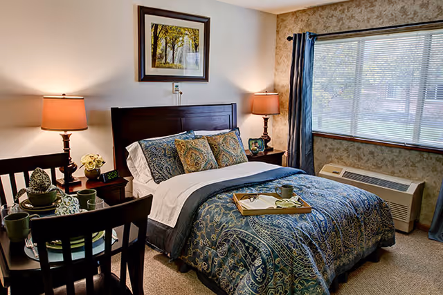 A cozy bedroom with a neatly made bed featuring a blue and green patterned comforter and multiple pillows. There are two bedside tables with lamps and decorative items. A framed picture hangs above the bed. A window with blue curtains lets in natural light, and a small dining table with chairs and place settings is visible in the foreground.