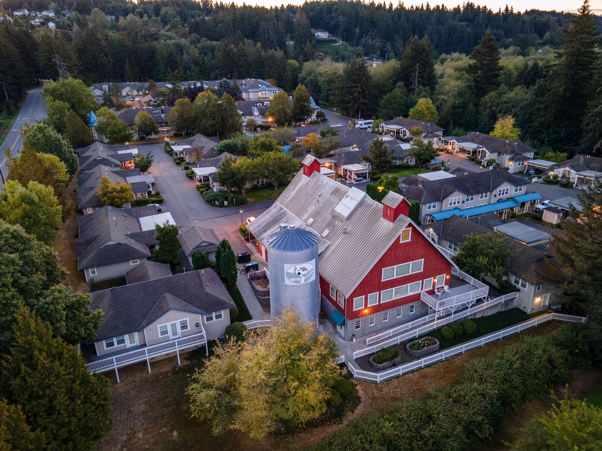 Aerial view of Country Meadows senior living campus centered on a red barn-style main building with a silo and surrounding residential cottages among trees.