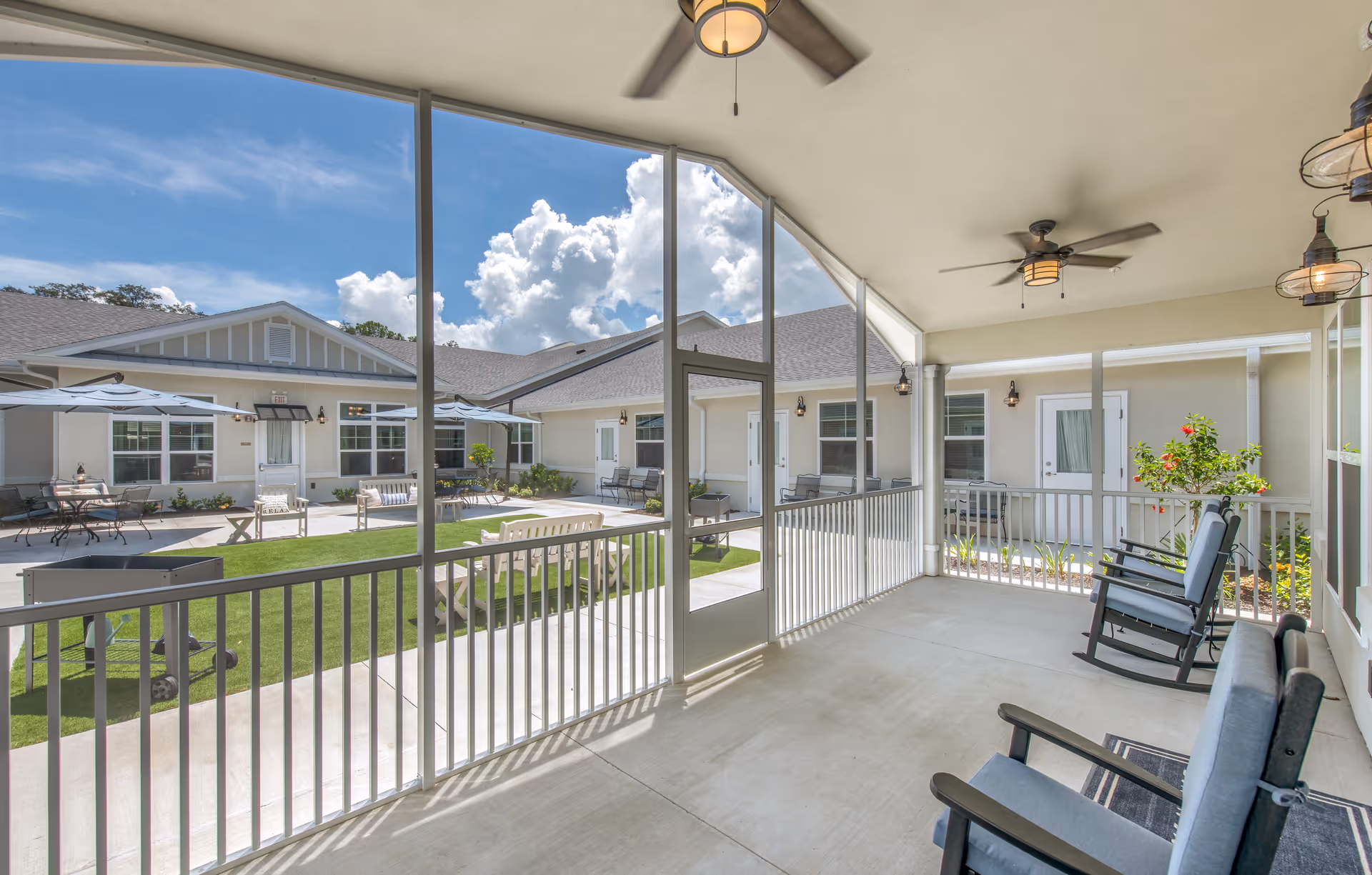 A screened-in porch area with ceiling fans and cushioned rocking chairs overlooking a courtyard with green artificial grass, benches, tables with umbrellas, and a barbecue grill. The courtyard is surrounded by single-story building structures under a bright blue sky with clouds.