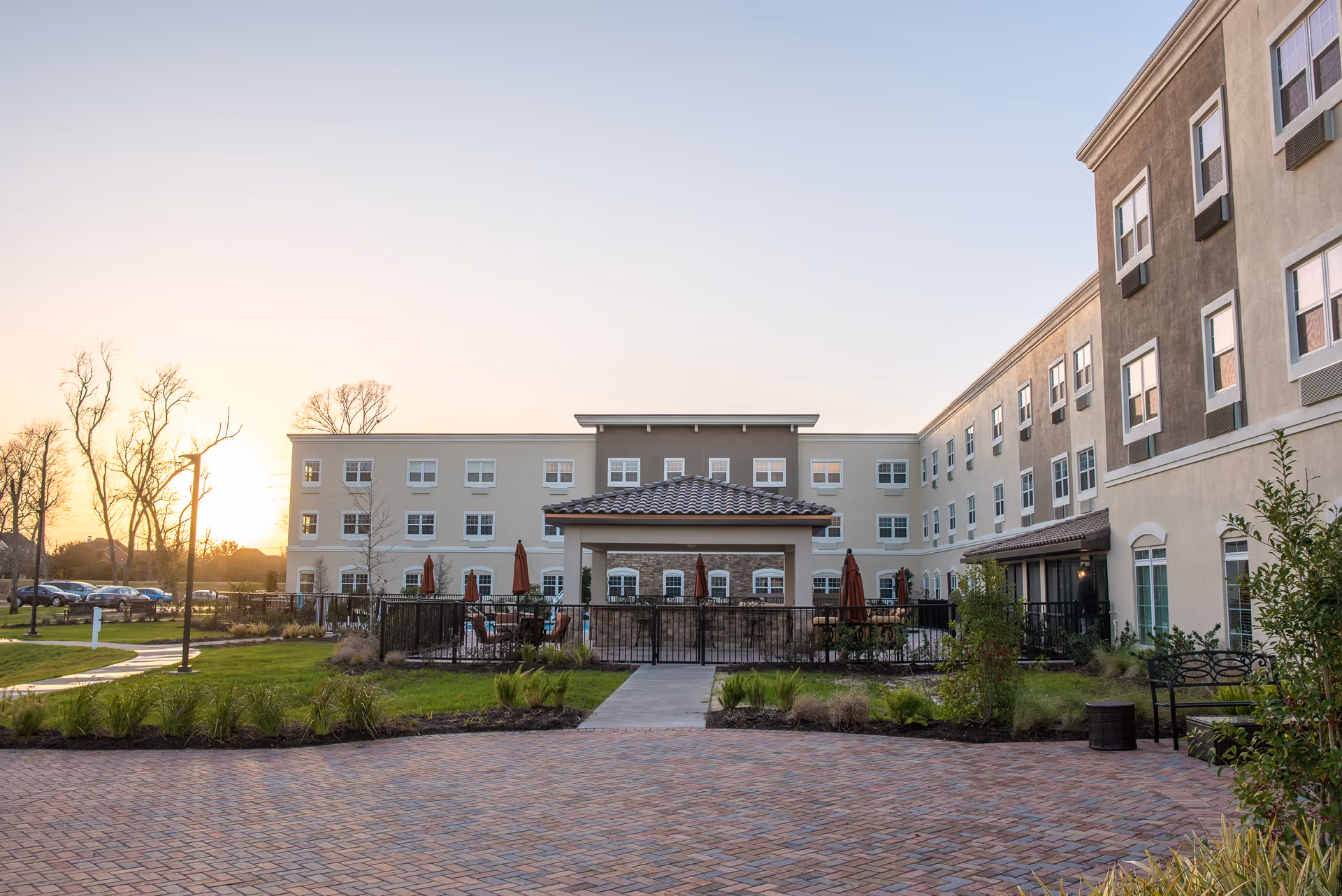 Exterior view of a senior living facility building at sunset with a paved courtyard, green lawn, and a covered outdoor seating area with tables and umbrellas.