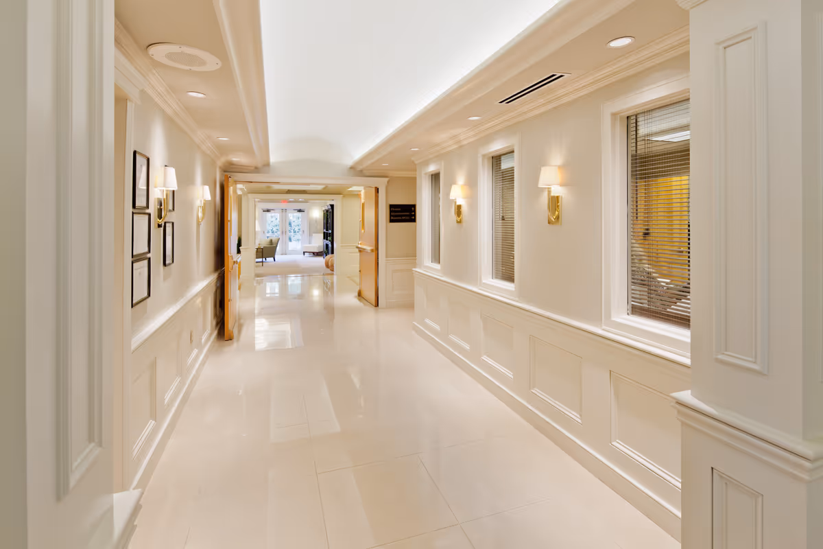 A bright, clean hallway in a senior living facility with cream-colored walls, recessed lighting, wall sconces, and framed pictures. The hallway leads to a seating area with chairs and large windows letting in natural light.