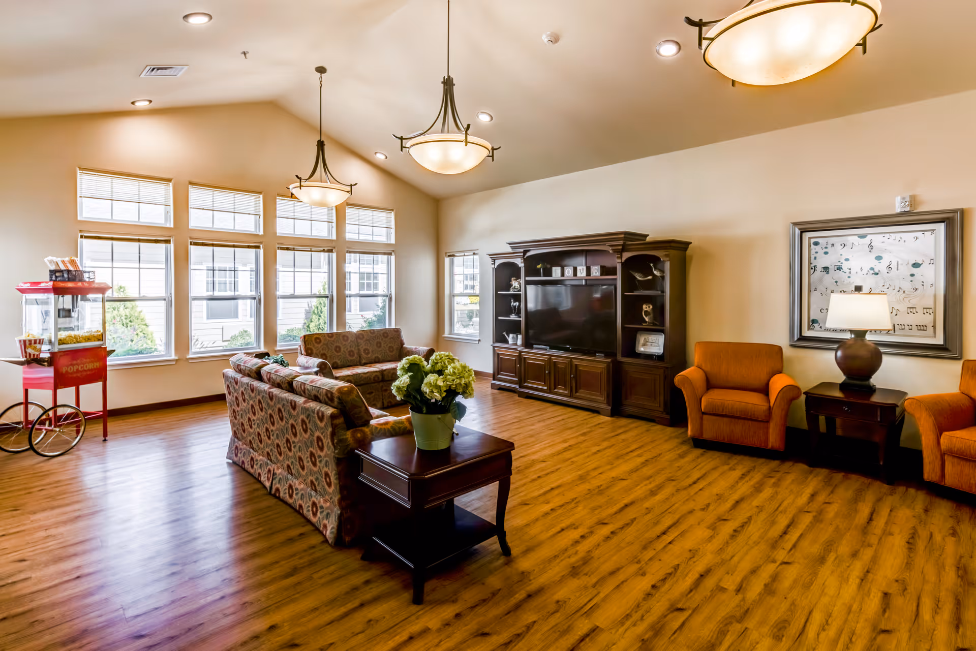 A spacious living room with wooden flooring, two patterned sofas, two orange armchairs, a dark wooden entertainment center with a TV, a side table with a lamp, a framed musical notes artwork on the wall, large windows letting in natural light, and a red popcorn machine in the corner.