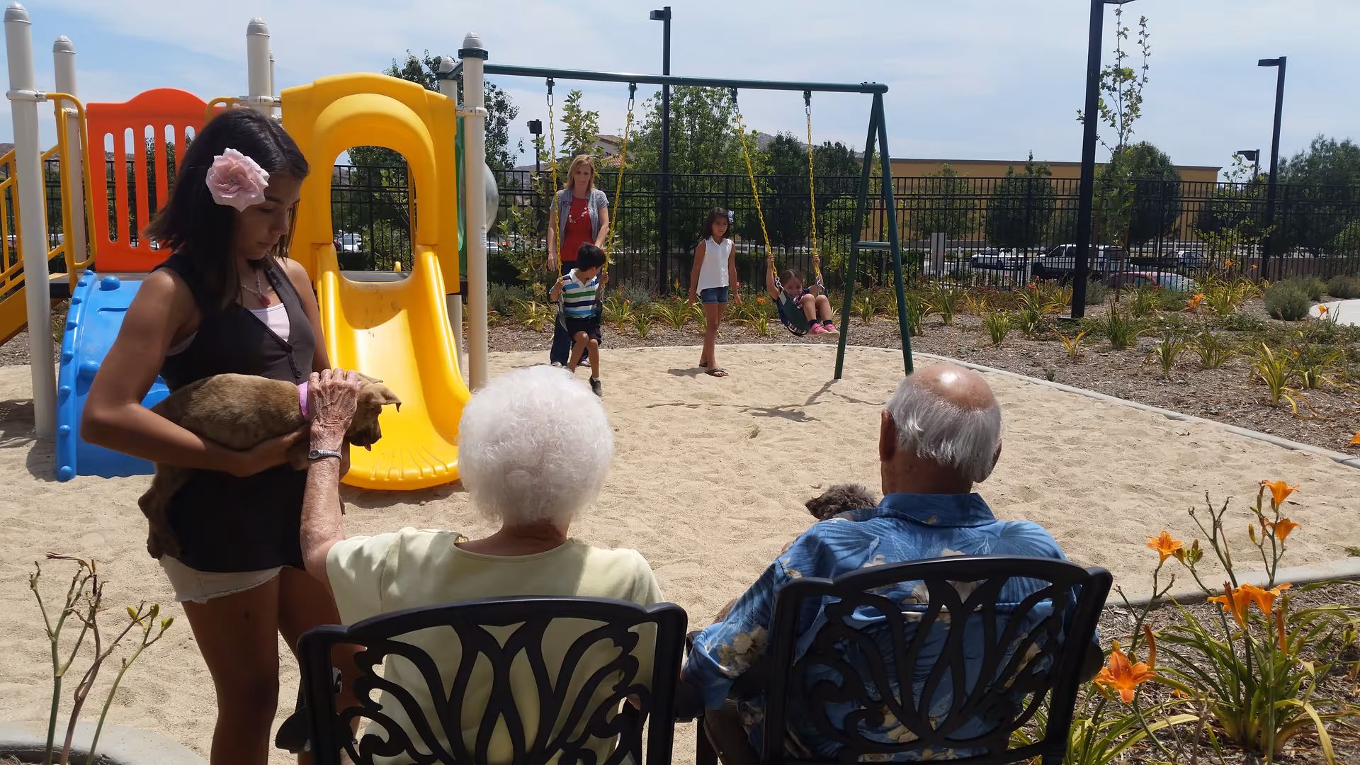 An outdoor playground area with a yellow slide and swings. Two elderly people are seated on black decorative chairs facing the playground. A young woman holding a small dog stands nearby, while children play on the swings and slide in the background. The area is surrounded by plants and a fence, with a clear sky overhead.