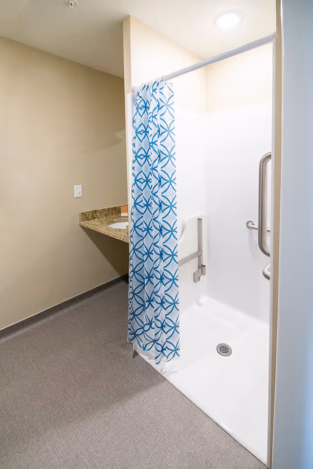 A clean, accessible shower area with a white shower base and walls, equipped with metal grab bars and a blue and white patterned shower curtain. Adjacent to the shower is a granite countertop with a sink, set against beige walls and a gray floor.