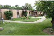 Single-story building with beige exterior walls and several windows, surrounded by well-maintained green grass, bushes, and trees. A paved walkway curves through the lawn leading to a covered patio area with outdoor seating.