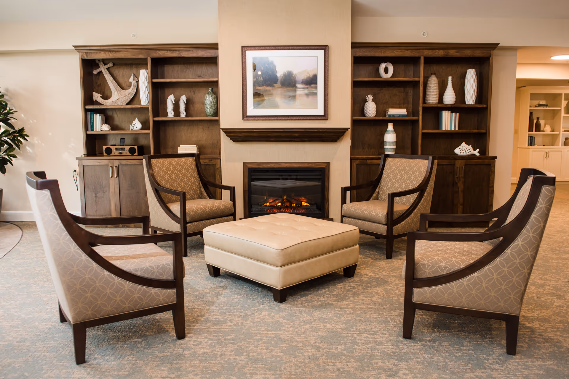 A cozy senior living common area with four patterned armchairs arranged around a large beige ottoman. Behind the seating is a fireplace with a framed landscape painting above it, flanked by wooden bookshelves decorated with various vases, books, and nautical-themed decor including an anchor and fish sculptures.