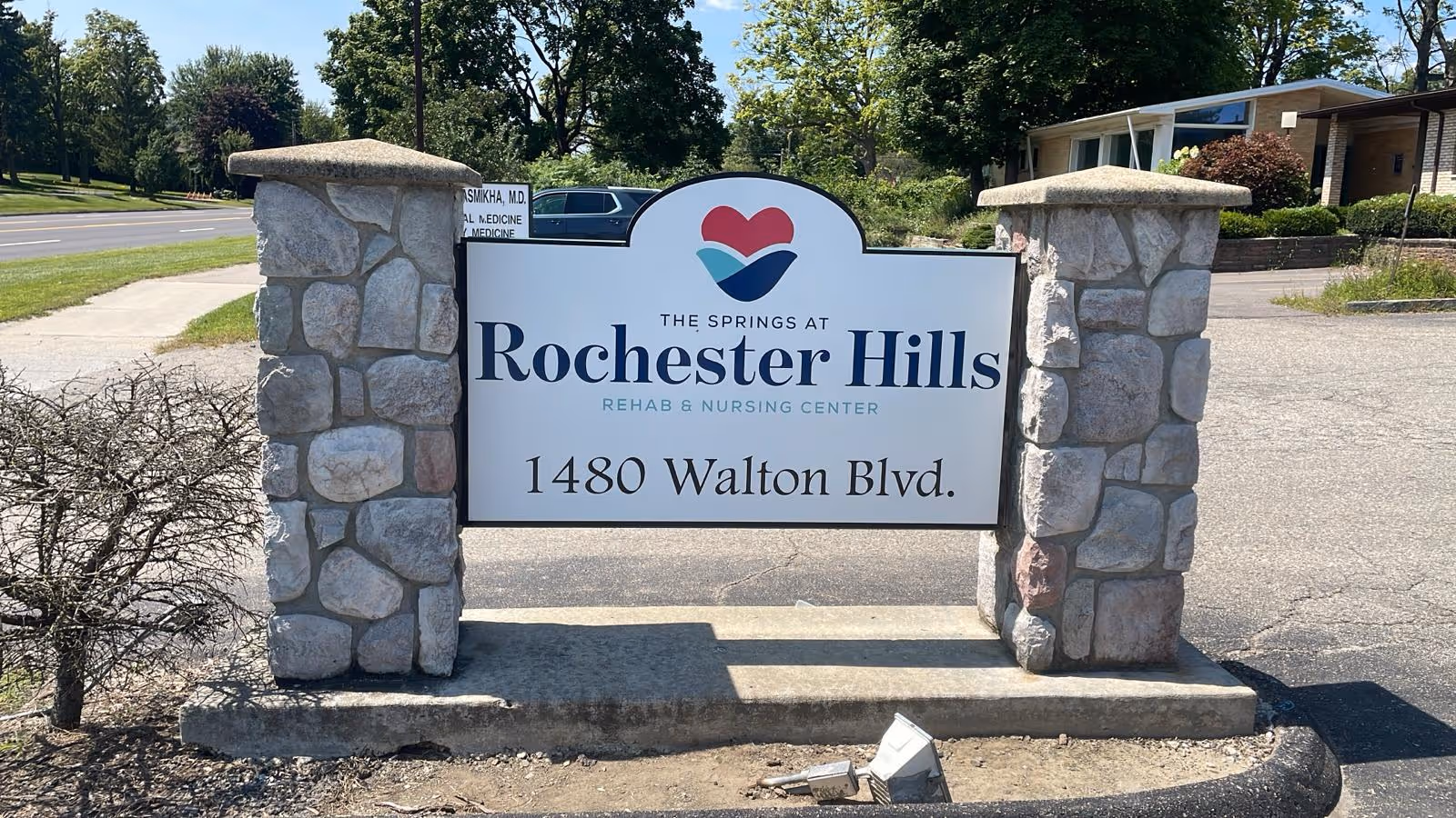 Outdoor stone sign for The Springs at Rochester Hills Rehab and Nursing Center located at 1480 Walton Blvd, with a road and trees in the background.