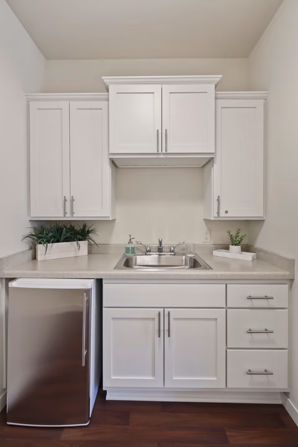 A small kitchen area with white cabinets, a stainless steel mini refrigerator, a stainless steel sink with a faucet, and a beige countertop. There are two small potted plants on the countertop, one on the left and one on the right side of the sink.