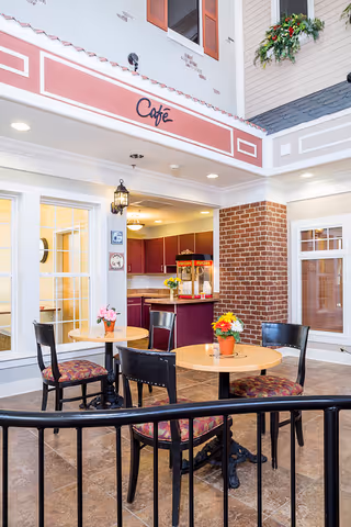 Indoor café area with round wooden tables and black chairs with patterned cushions. Each table has a small flower pot with colorful flowers. The café counter in the background has a popcorn machine and a vase with yellow flowers. The walls are decorated with brick and light-colored paneling, and there are large windows and a sign above the café counter that reads 'Café'.