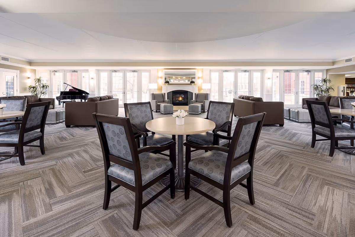 A spacious and well-lit common area in a senior living facility featuring several round tables with four cushioned chairs each. In the background, there are comfortable sofas and armchairs arranged around a fireplace with a mirror above it. Large windows and glass doors let in natural light, and a grand piano is visible on the left side of the room. The room has a modern carpeted floor and neutral-colored walls.