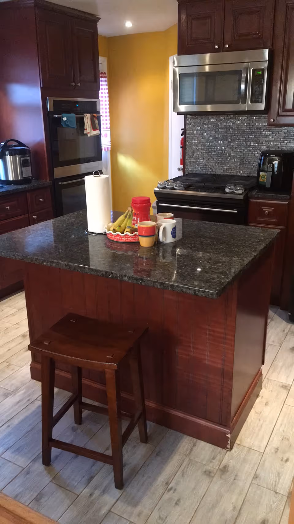 A kitchen with dark wood cabinets and a granite countertop island. On the island are a paper towel holder, a bowl of bananas, a red container, and three coffee mugs. There is a wooden stool in front of the island. The kitchen features a stainless steel microwave above a stove with a tiled backsplash, and a yellow wall in the background.